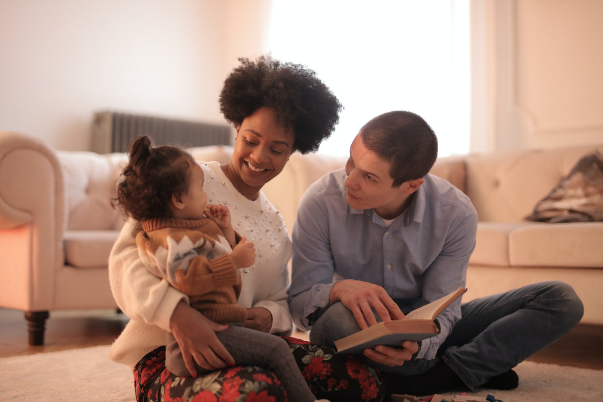 Two young parents sit on their living room floor. Mom holds a young infant in her arms and dad is reading to her from a book.